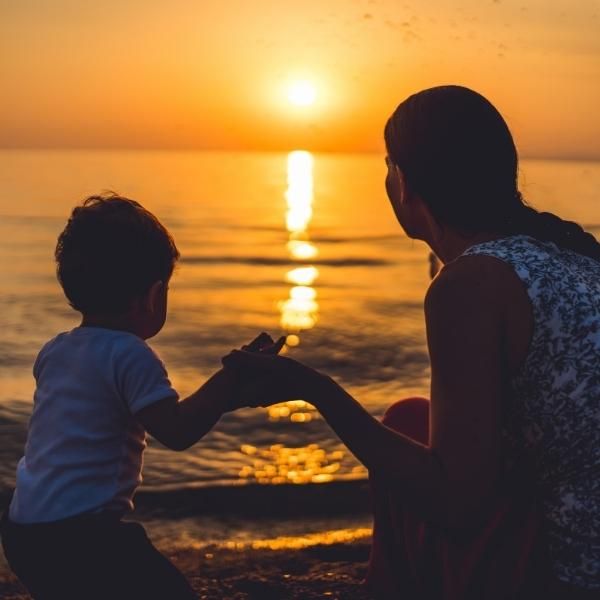 Foto de una madre y un hijo mirando una puesta de Sol desde la playa.