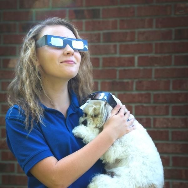 Foto de una mujer con gafas protectoras para mirar el Sol, mientras sostiene un perro en sus brazos.