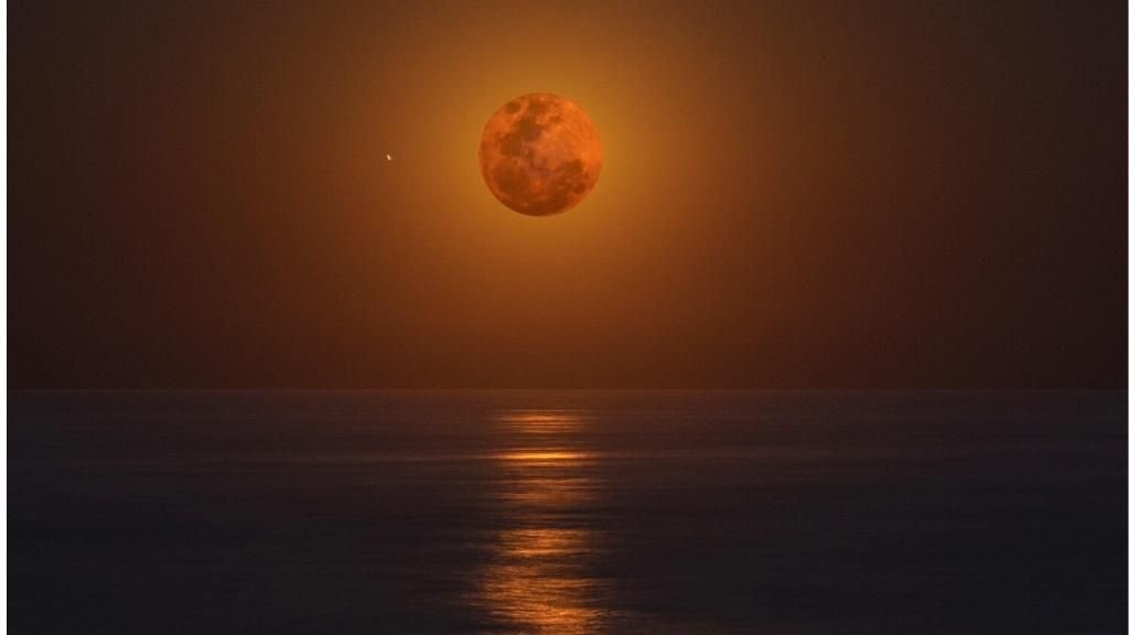 Foto de la Luna llena al fondo rojiza y sobre el mar en el horizonte.